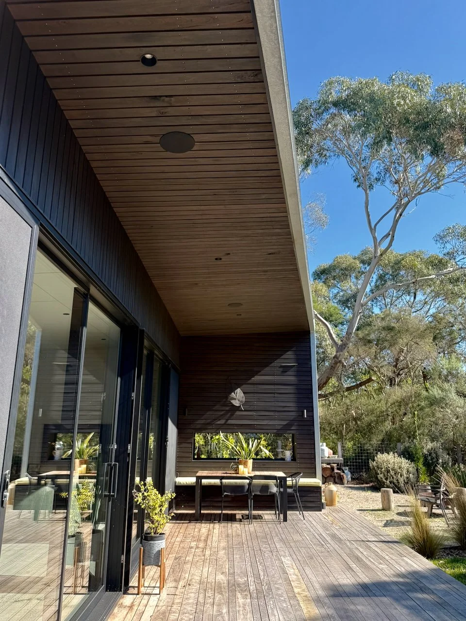 Modern house exterior with wooden deck, large glass sliding doors, potted plants, outdoor table with chairs, and trees in the background under a clear blue sky.