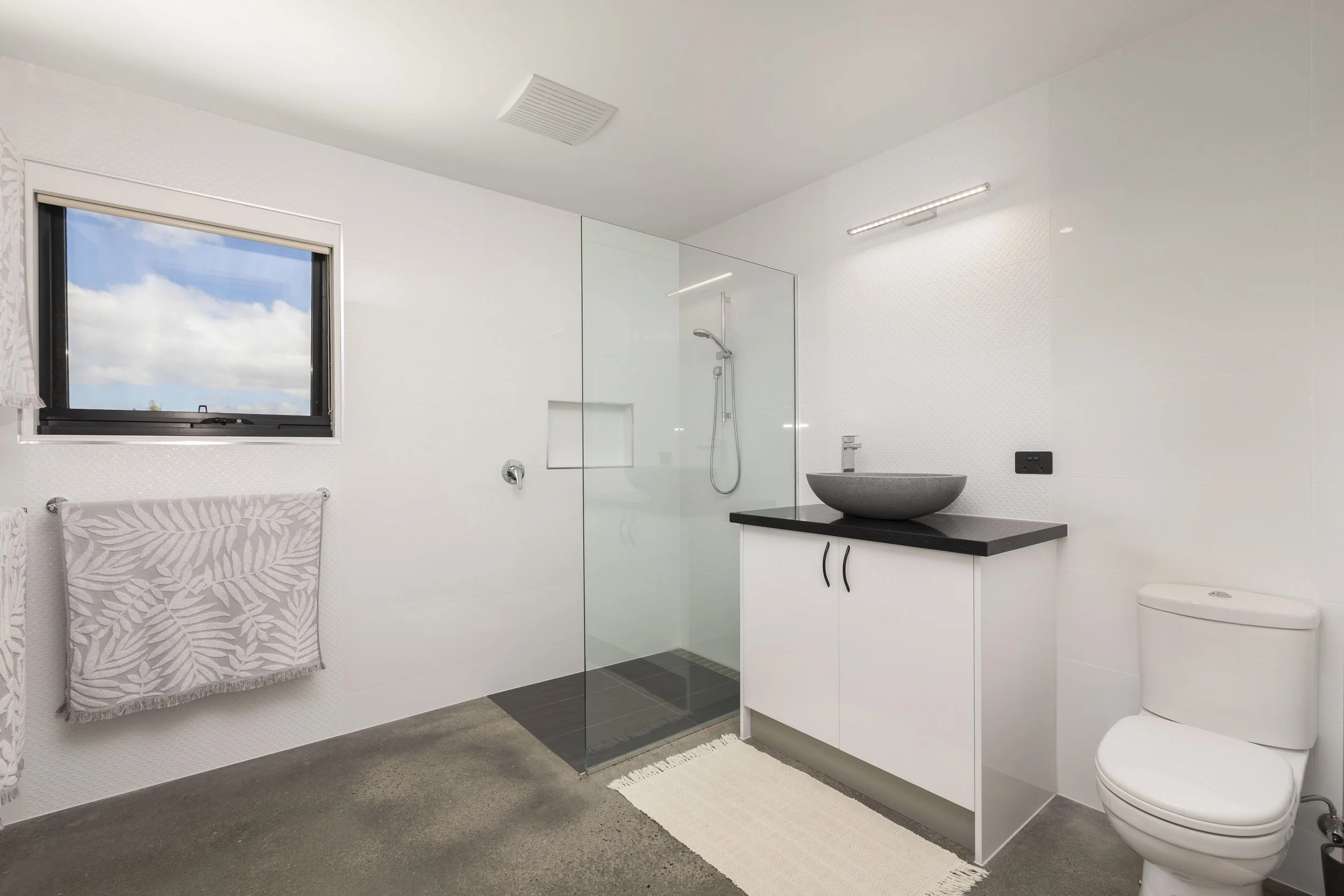 Modern bathroom with a window showing a blue sky and clouds, a towel hanging on a wall, a shower with glass partition, a white vanity with a vessel sink, and a toilet. The walls are white with textured tile, and the floor is gray.