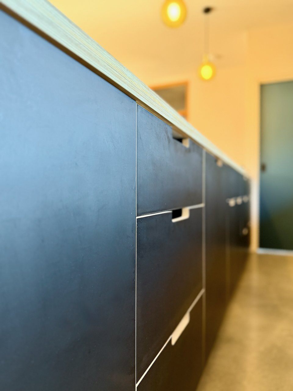 Close-up of a dark-colored cabinet with sleek handles and a wooden countertop, viewed from a low angle with warm lighting in the background.