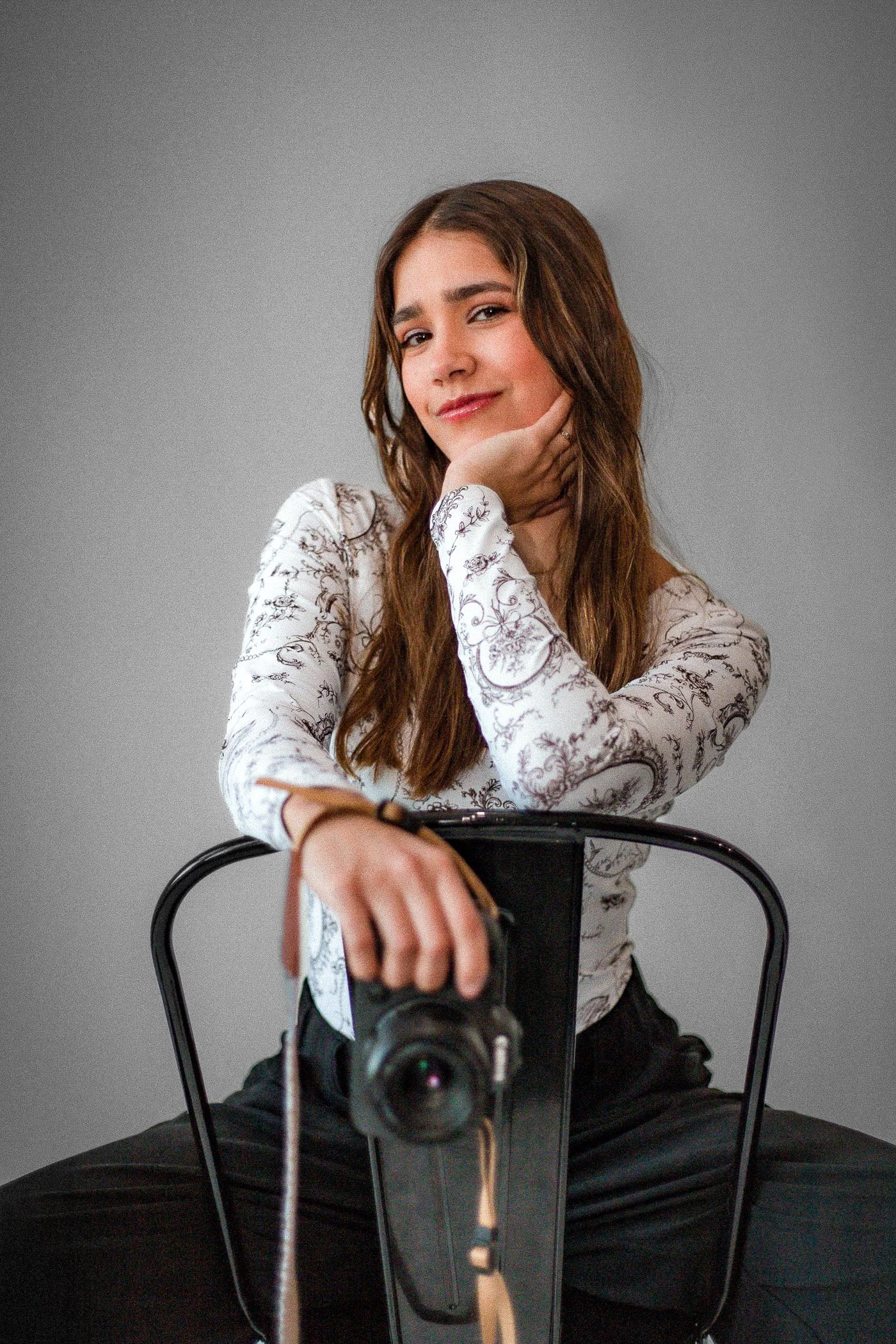 A young woman with long brown hair sitting on a black chair in front of a gray background. She is wearing a long-sleeve shirt with a floral pattern and black pants. She is looking at the camera with a subtle smile, resting her chin on her right hand while reaching towards the camera with her left hand, which is attached to a camera on a tripod or stand.