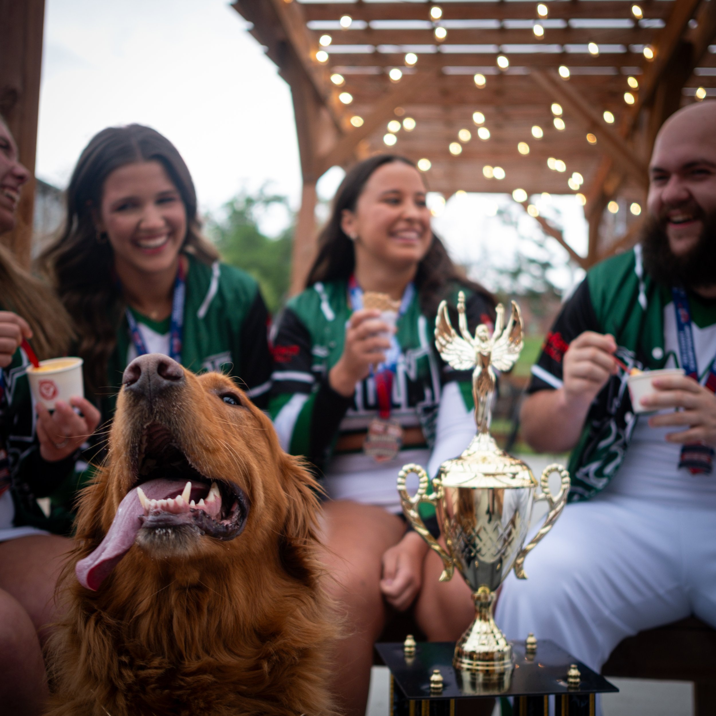 People at a celebration or competition with a dog in the foreground and a chess trophy on a table in the background.