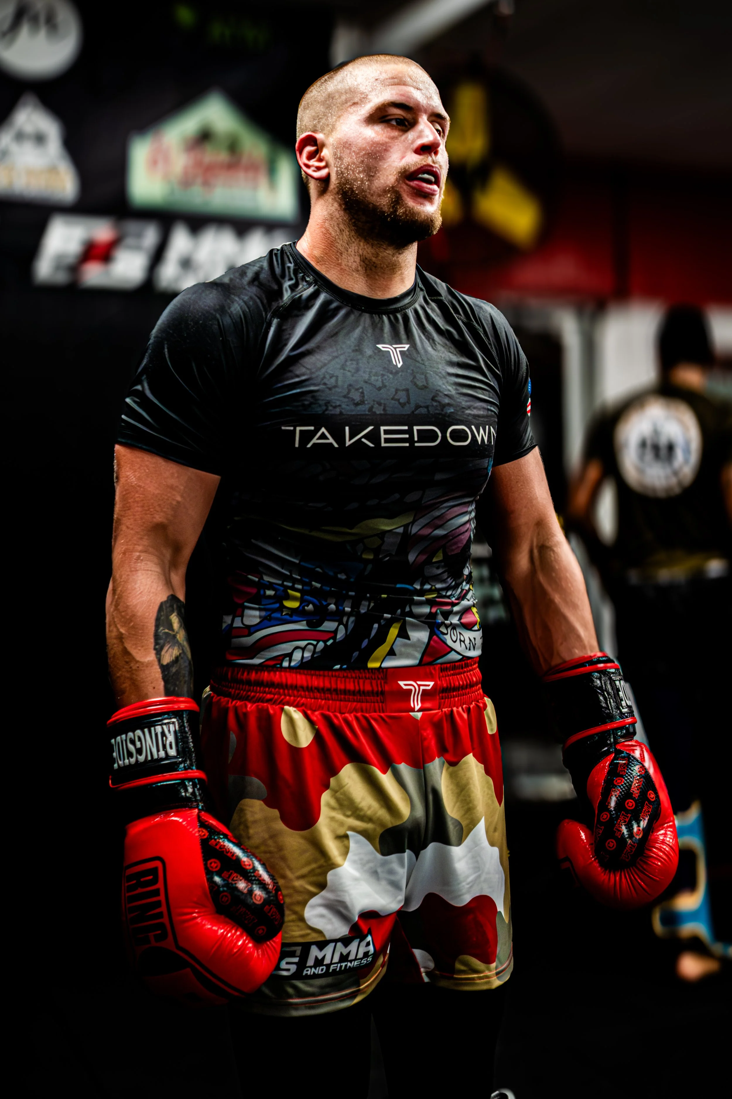 A male boxer in red gloves and colorful shorts stands in a gym, wearing a black Takedown Fitness shirt, with a focused expression.