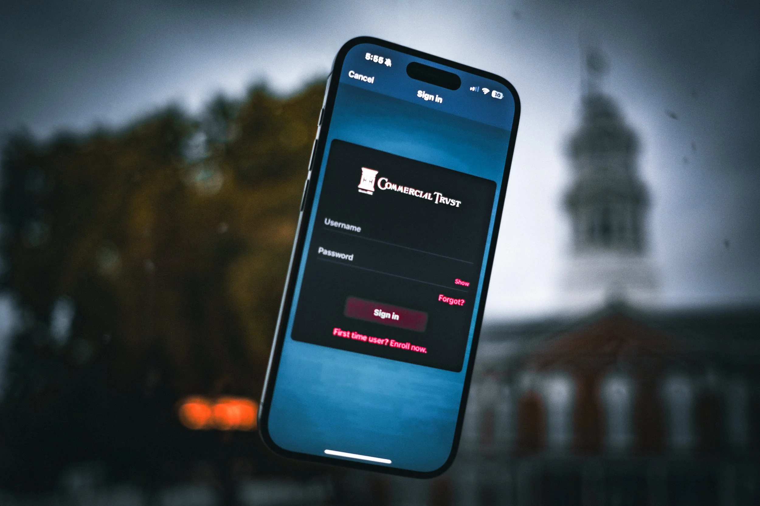 A smartphone displaying a login screen for a service called 'Commercial Trust,' with fields for username and password, and options for sign-in, forgot password, and new user enrollment. The phone is held in front of a blurred outdoor background during dusk or evening.