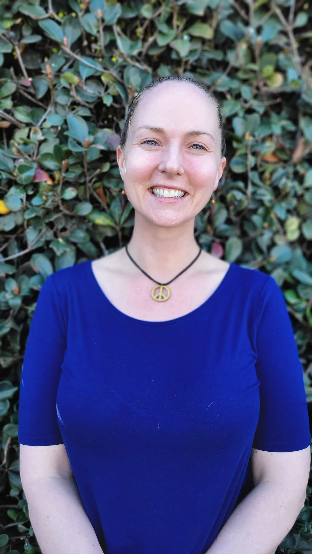 Smiling woman with a peace sign necklace, wearing a blue top, standing in front of a leafy green background.