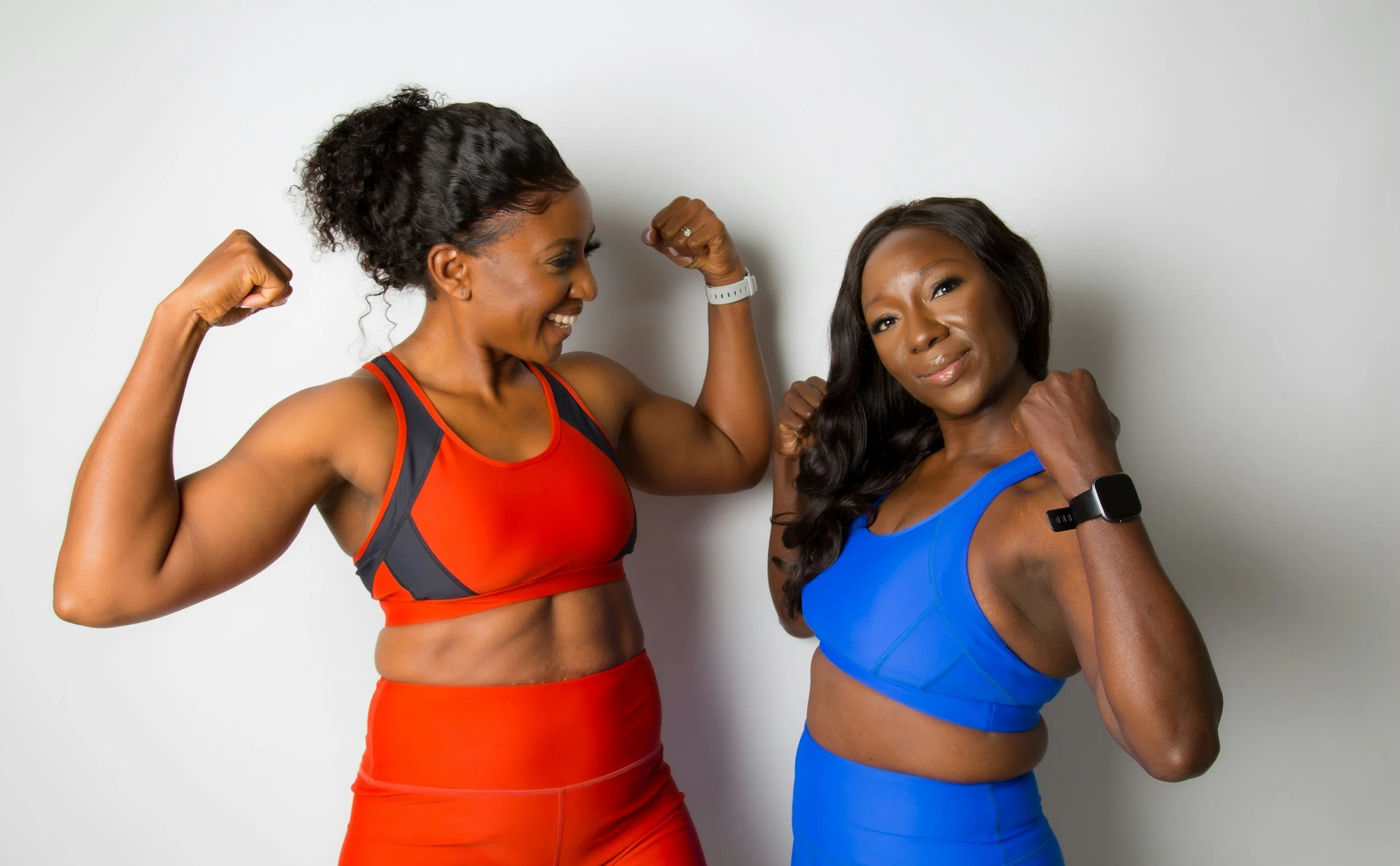 Two women in athletic clothing, flexing their muscles and smiling at each other against a white background.