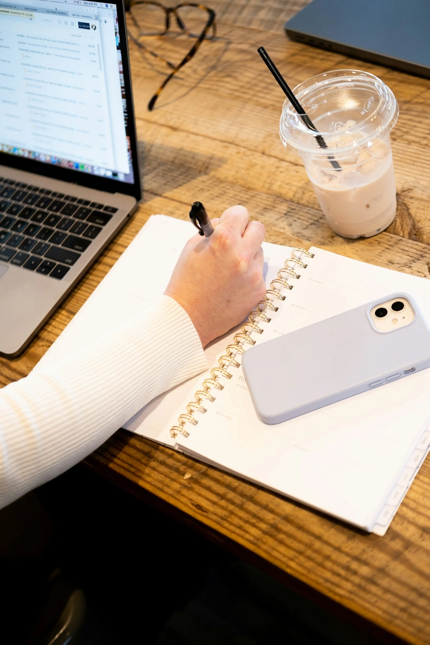 Overhead view of a person writing in a planner with a black pen, on a wooden table, alongside a silver laptop, glasses, a clear plastic cup with iced coffee, a closed laptop, and a white smartphone.