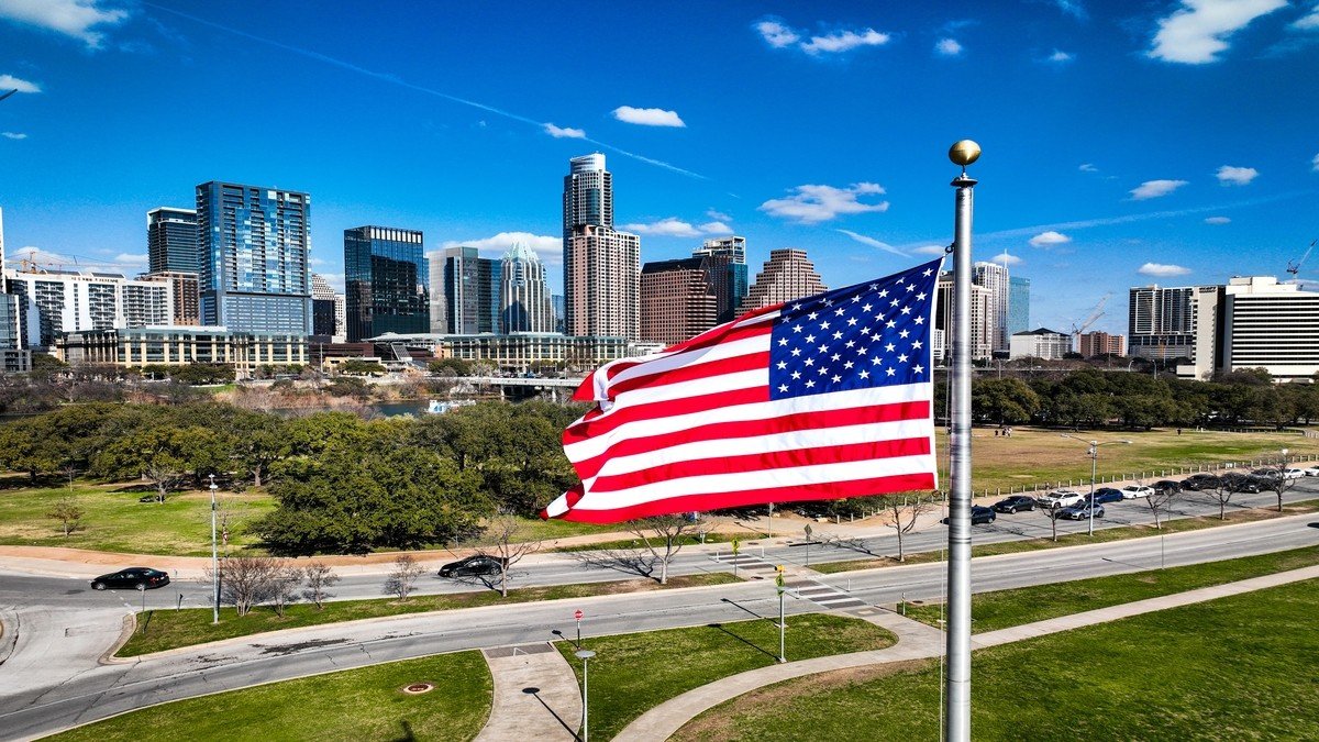 Bandeira dos Estados Unidos tremulando ao vento em frente ao skyline de edifícios de uma cidade, com áreas verdes e céu azul ao fundo.