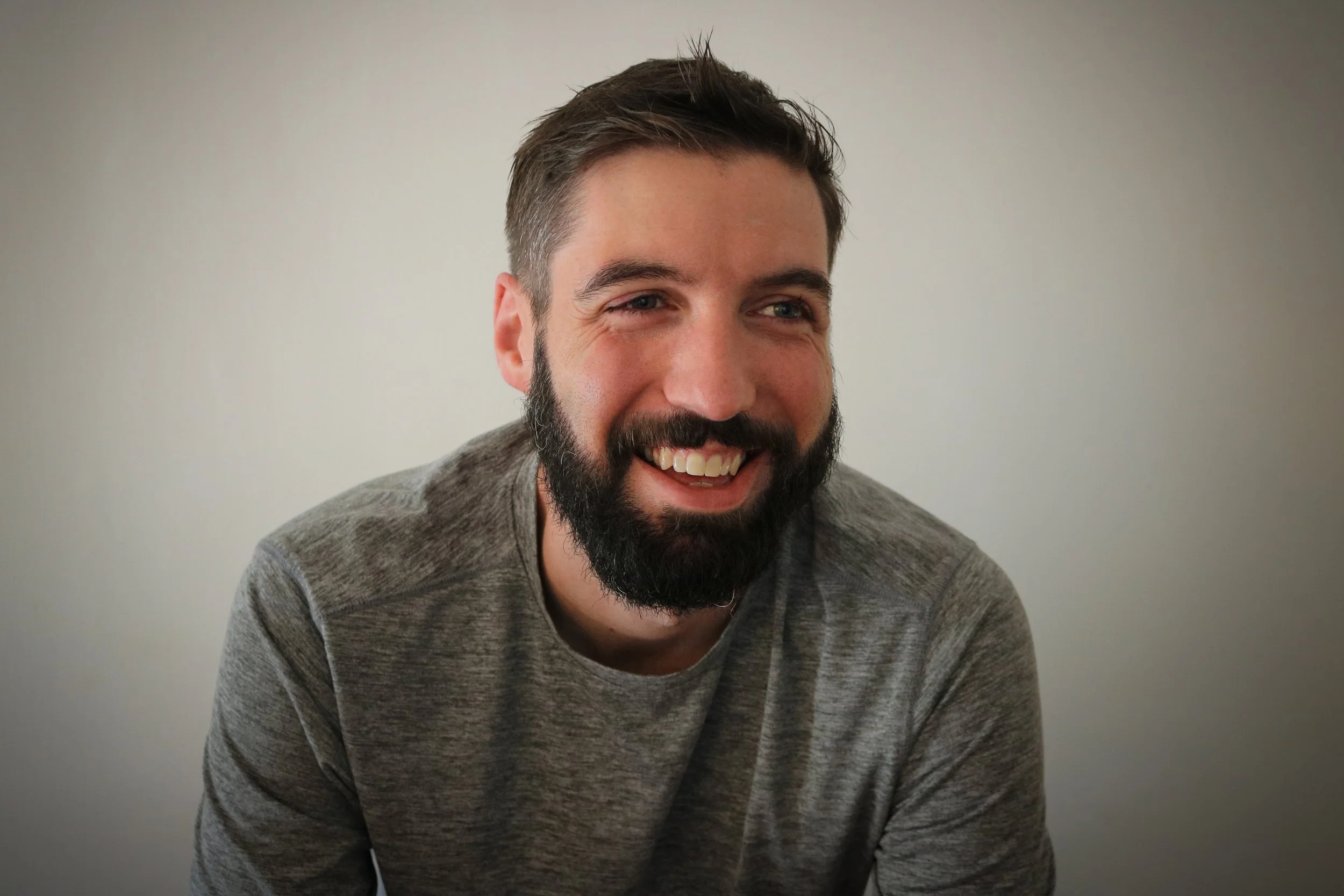 Smiling man with dark hair and beard in gray shirt, looking to the side against a plain white wall.