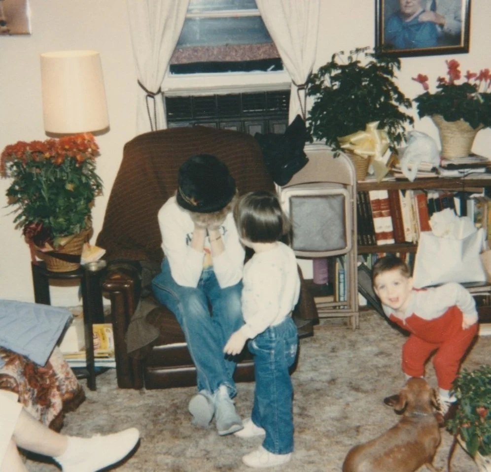 A woman with two children sits on a chair in a cozy living room decorated with plants, bookshelves, and family photos. One child stands in front of her and the other leans in, smiling. A small dog is on the carpet, and the room has a window with curtains.