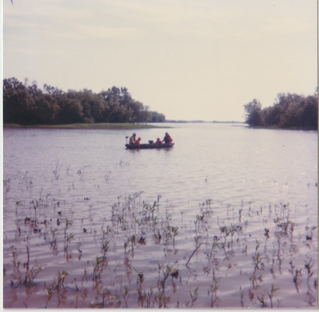 People fishing from a boat on a calm river with trees along the shoreline in the background.