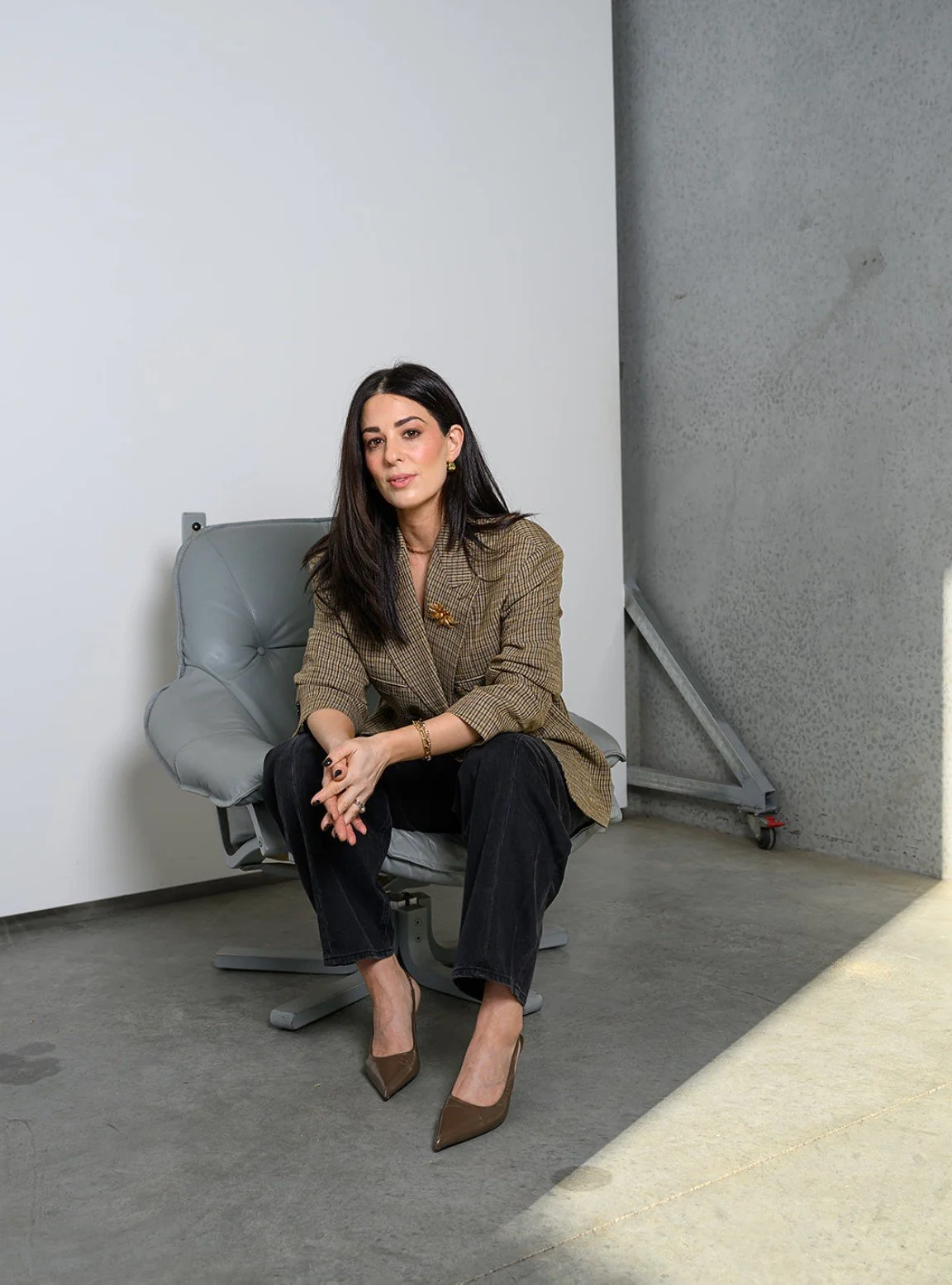 Woman sitting on a gray cushioned chair in a modern space with gray walls, dressed in a brown checked blazer, black pants, and heels, with one hand resting on her knee and the other on her lap.
