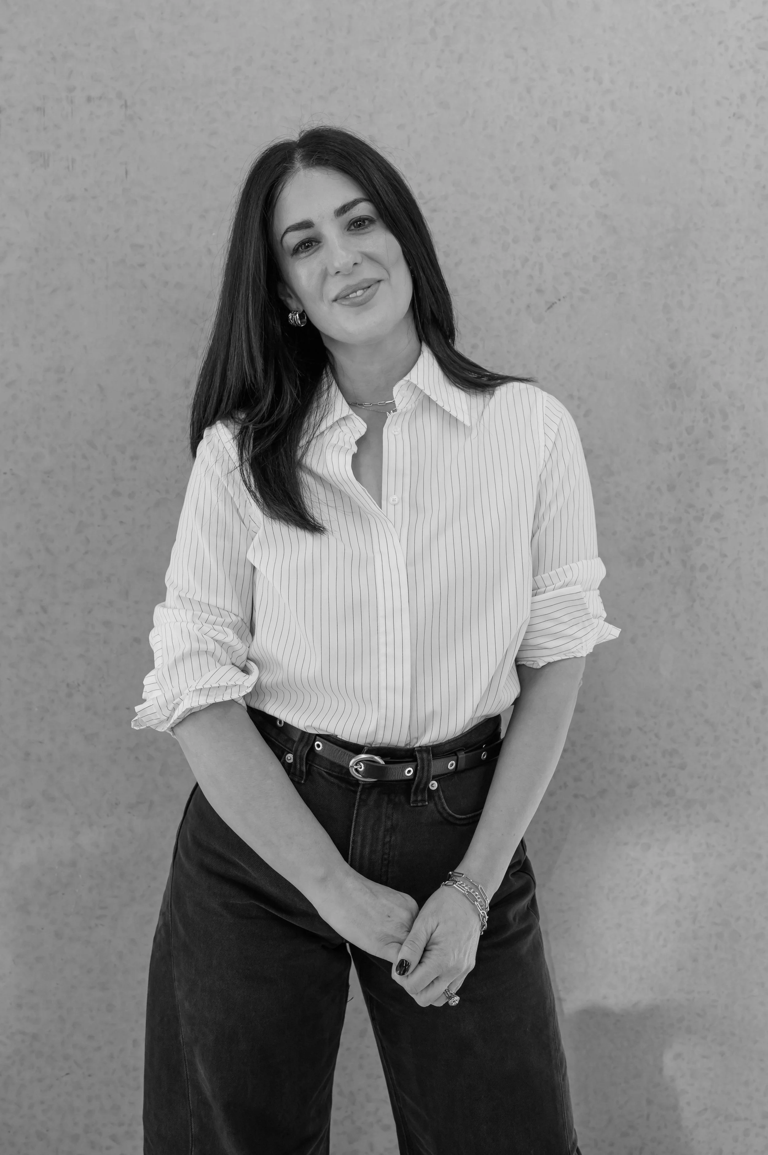 A woman with dark hair, wearing a striped button-down shirt and dark jeans, standing in front of a textured wall, smiling slightly with her arms crossed.