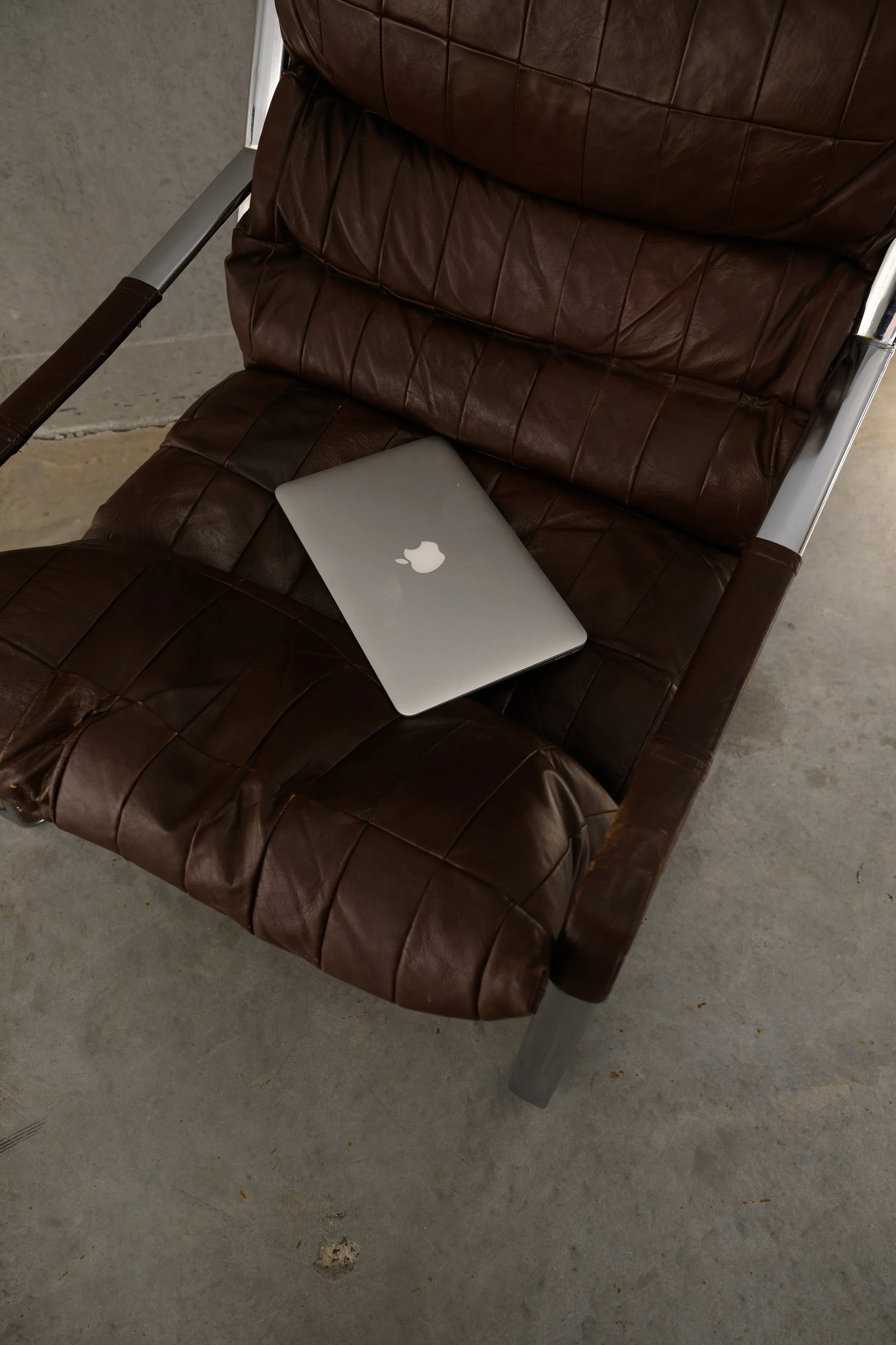 A brown leather chair with metal armrests and legs, with a closed silver MacBook laptop placed on the seat. The chair is positioned on a gray concrete floor.