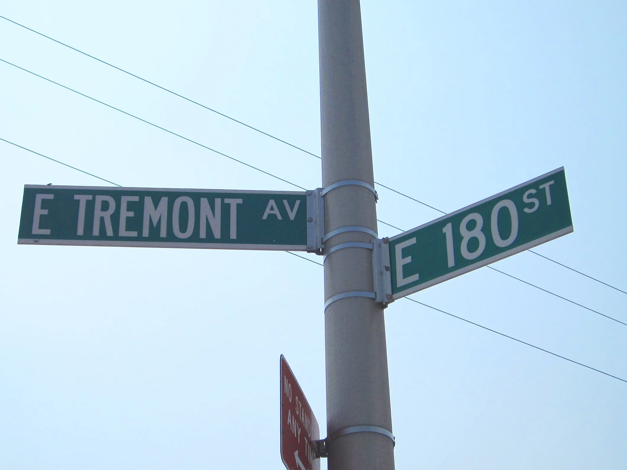 Street signs at an intersection of E Tremont Ave and E 180 St, with a utility pole and power lines against a light blue sky.
