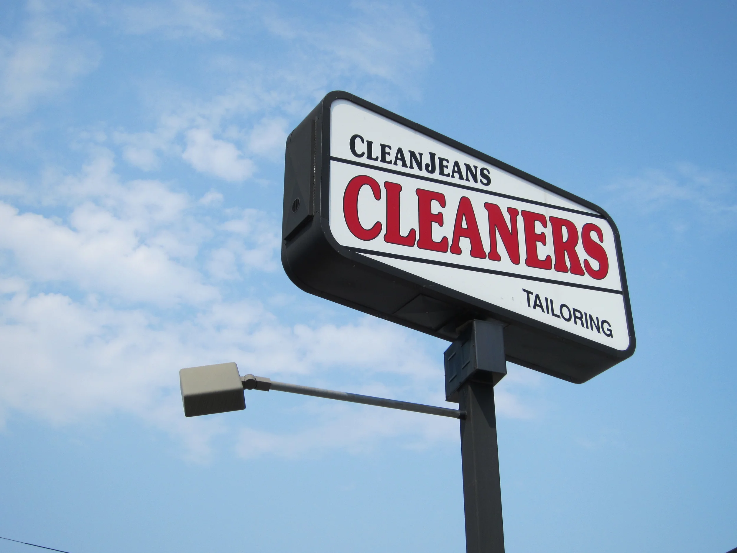 A roadside sign that reads 'CLEANJEANS' at the top and 'CLEANERS' below, with additional text that is partially visible. The sign is mounted on a pole against a blue sky with some clouds.