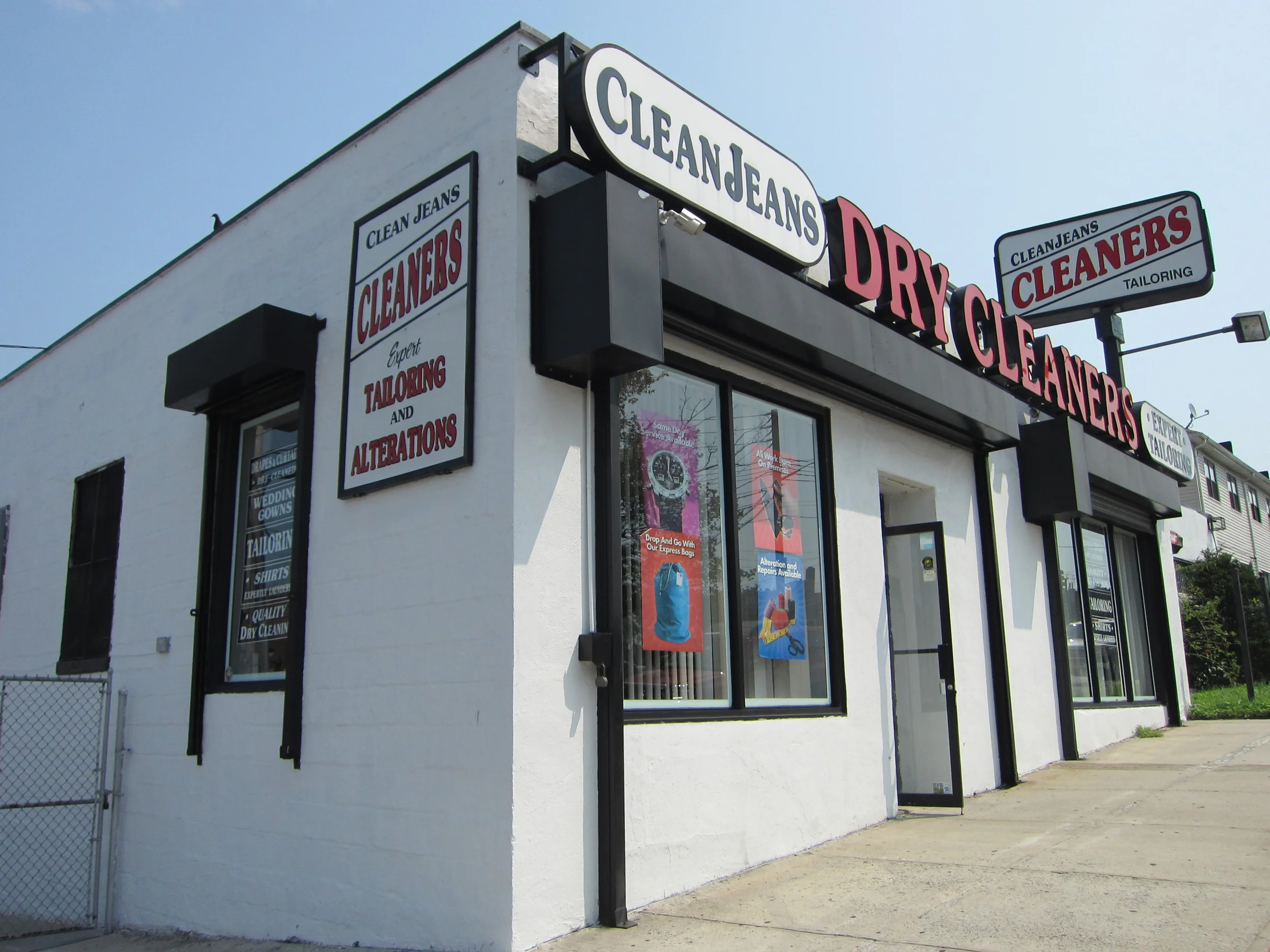 Corner of a white building with black trim and multiple signs reading 'Clean Jeans' and 'Cleaners,' with notices about tailoring and alterations. The building has large windows, and there is a narrow sidewalk in front.