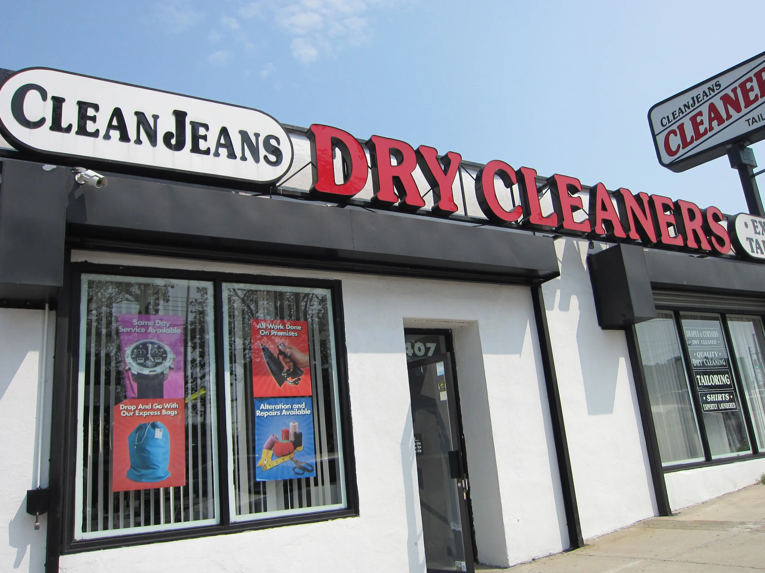 Exterior of a dry cleaner shop with signs indicating services such as cleaning and repair, and posters advertising drop-off bags, repair services, and tailoring.