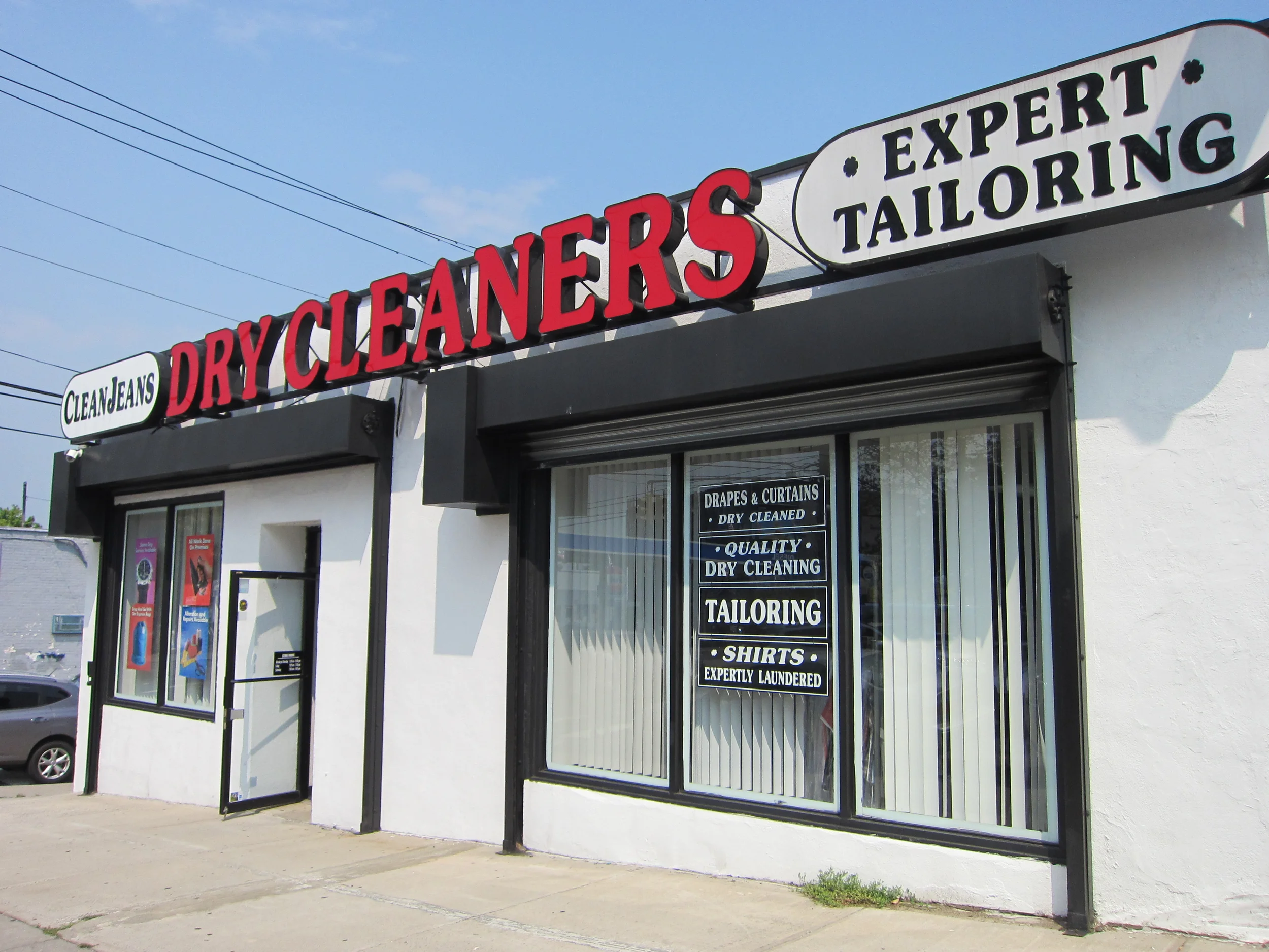 Front of a dry cleaner and tailoring shop with a sign that reads 'Dry Cleaners' in red letters and 'Expert Tailoring' in black letters on a white background. The shop has large windows displaying various signs for services including drapes and curtai