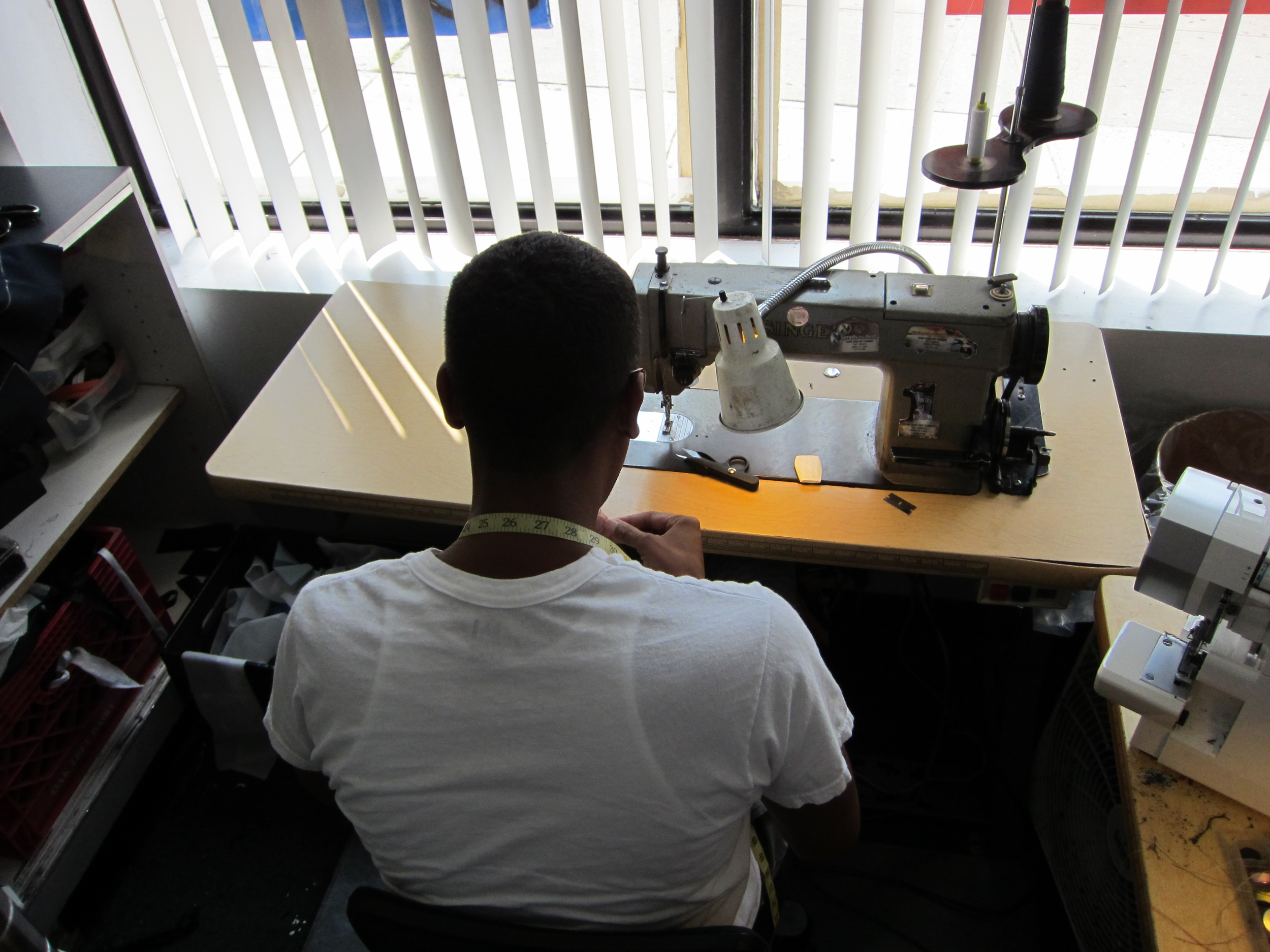 Person sitting at a work desk sewing on a sewing machine in a room with white window blinds.