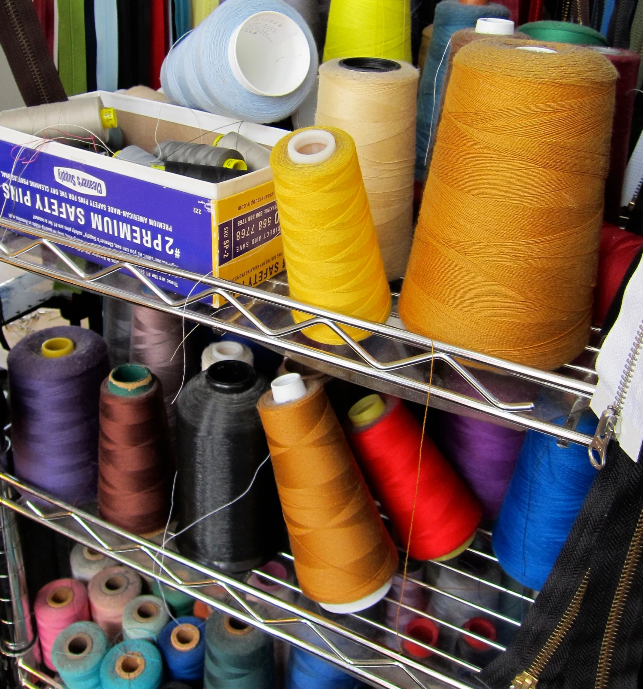 Spools of colorful thread in yellow, orange, red, green, blue, purple, grey, black, white, and pink on a metal rack, with a box of sewing pins and a black zipper in the background.