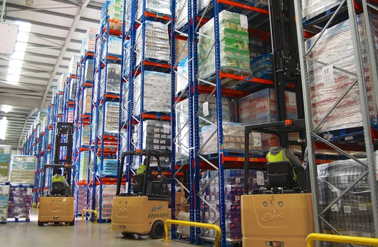 Warehouse with tall shelves stocked with pallets of goods, forklifts moving through the aisles, and workers operating the equipment.