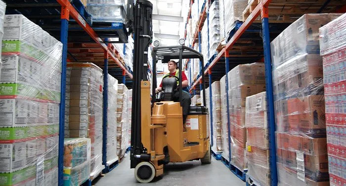 Person operating a forklift in a warehouse aisle filled with stacked boxes on shelves.