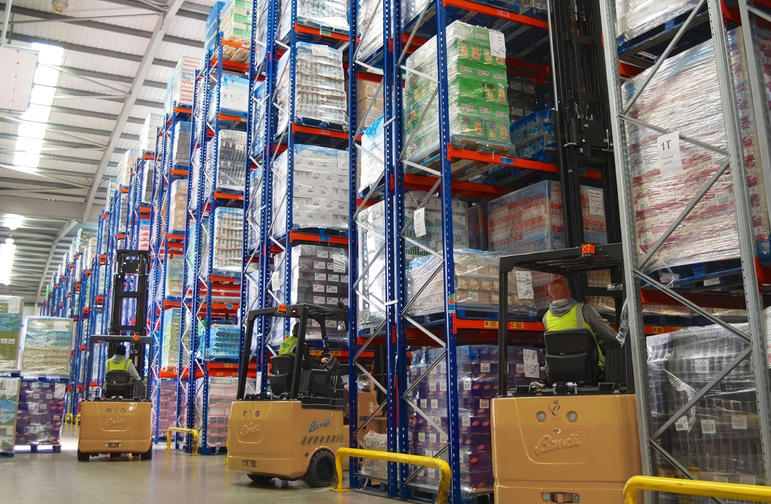 Inside a warehouse with tall blue metal storage racks holding various boxed products. Several workers in yellow vests are operating pallet jacks to move pallets of merchandise.