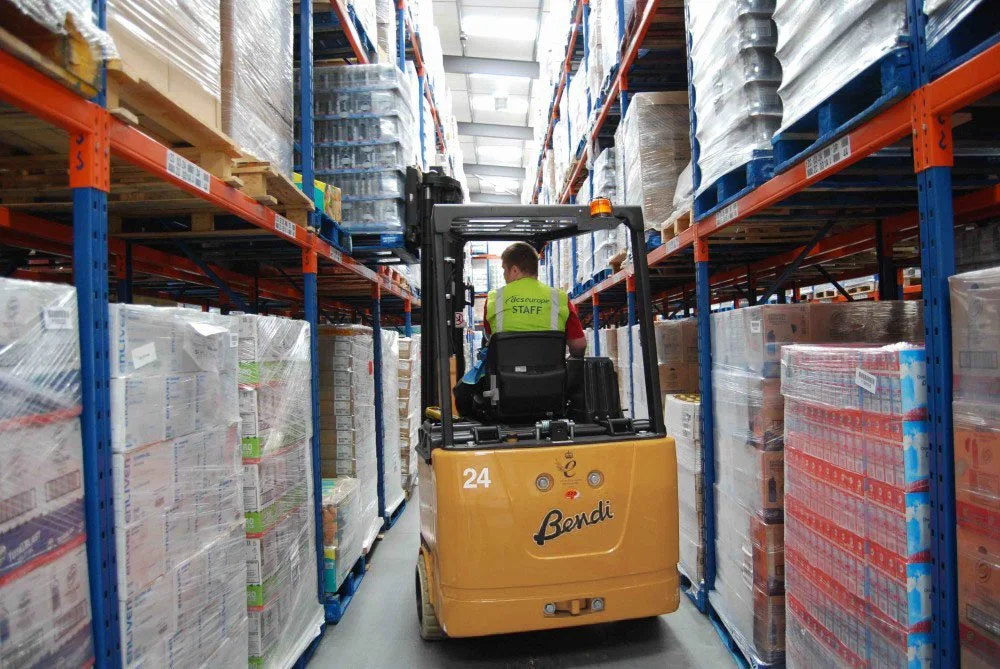 Warehouse aisle with shelves stocked with products wrapped in plastic; forklift operator with a green safety vest labeled 'STAFF'.