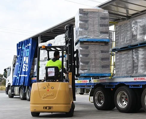 A person operating a forklift truck is moving large wrapped pallets of goods on a truck bed, with a blue curtain side truck in the background.