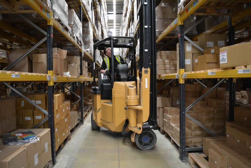 A worker operates a forklift in a warehouse aisle surrounded by shelves stacked with brown cardboard boxes.
