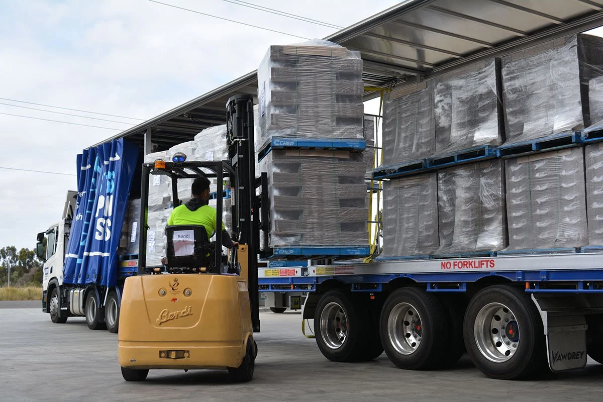 A forklift operator in a high-visibility shirt is lifting pallets onto a flatbed truck that is loaded with large boxed items wrapped in plastic. The truck has a sign that says 'No Forklifts' on the side.