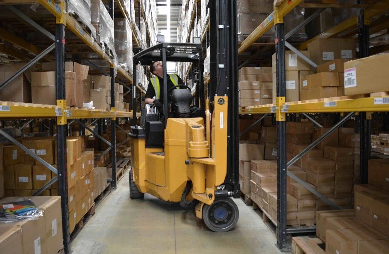 Warehouse worker operating a forklift among shelves filled with boxes and packages.