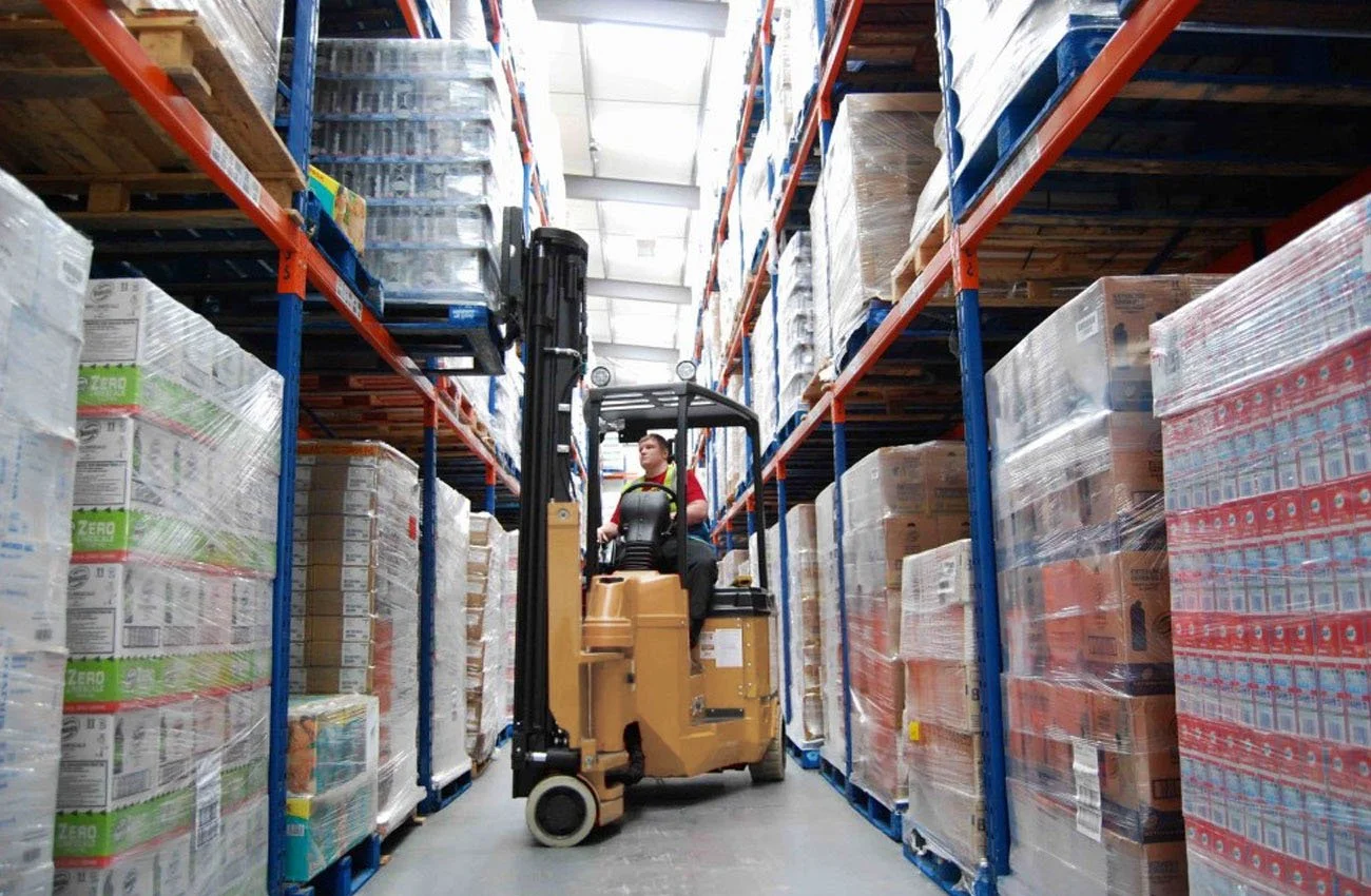 A worker operating a forklift inside a warehouse aisle, surrounded by shelves stacked with pallets of goods wrapped in plastic.