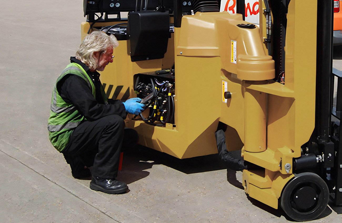 A man in a black shirt, black pants, and a green safety vest kneeling on the ground, working on a yellow industrial machine with an electronic device in his hands.