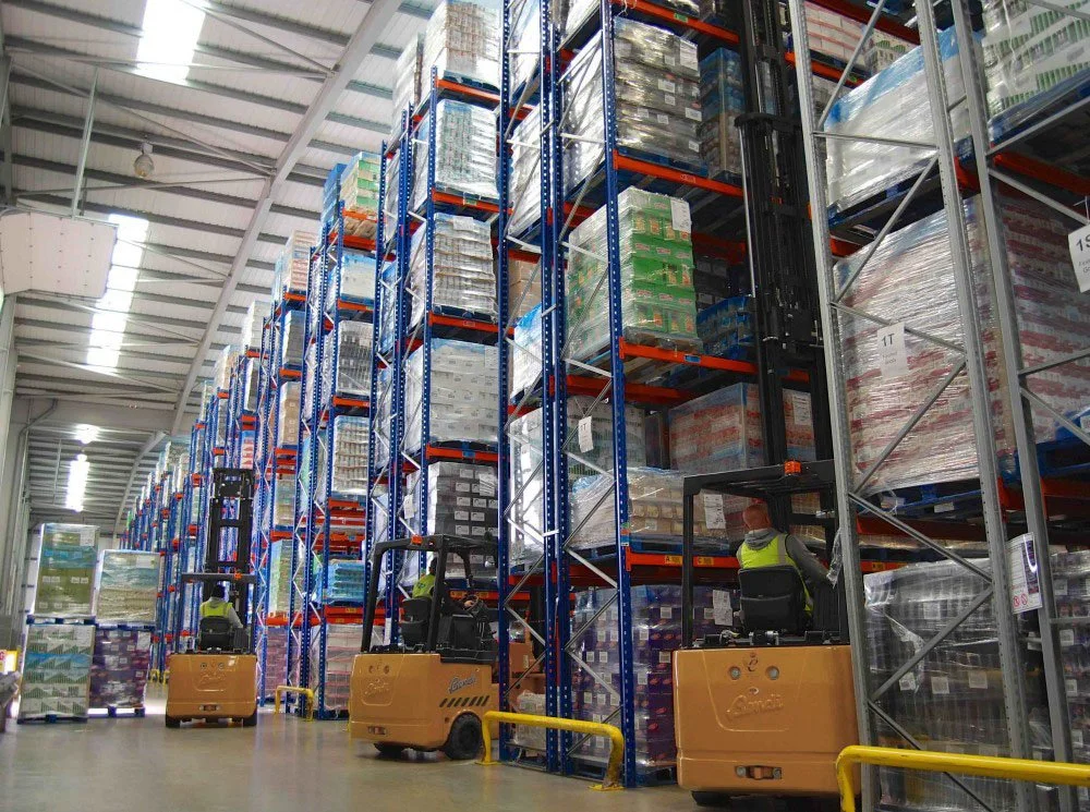 Warehouse with tall shelves stacked with boxes and pallets, workers operating forklifts.