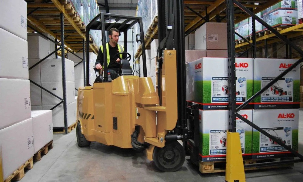 A worker operating a yellow forklift in a warehouse, moving boxes of AL-KO products on a shelf.