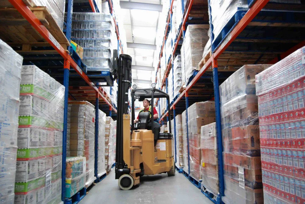 A person operating a forklift in a warehouse aisle with high shelves stacked with boxed and palletized goods.