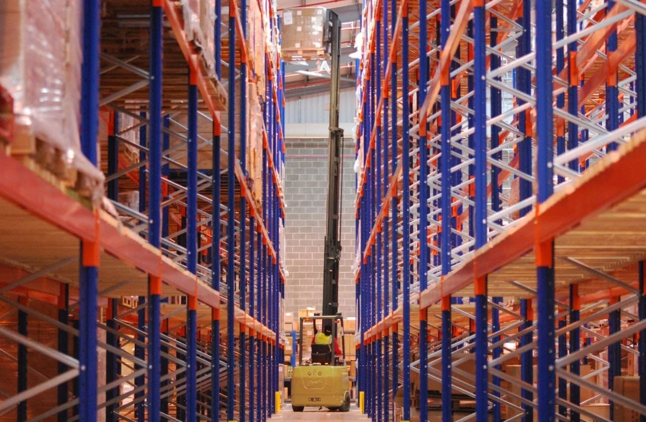 Inside a large warehouse with tall metal shelving units, a forklift with an extended lifting arm is in the center, stocking or retrieving pallets.