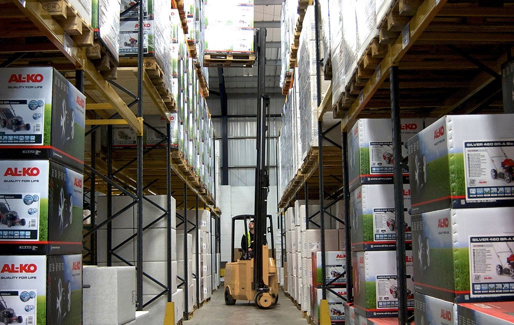 Warehouse aisle with tall shelves stocked with boxed products, and a forklift operated by a worker in a high-visibility vest moving through the aisle.