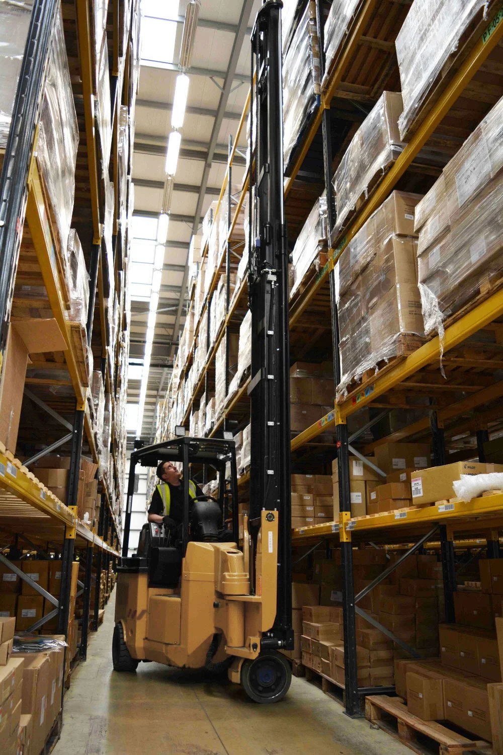A warehouse worker operating a forklift to reach high shelves filled with boxes and pallets.