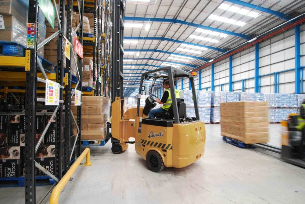A worker operating a yellow forklift inside a large warehouse, moving a pallet of products on a yellow-handled vehicle with shelves filled with boxed items in the background.
