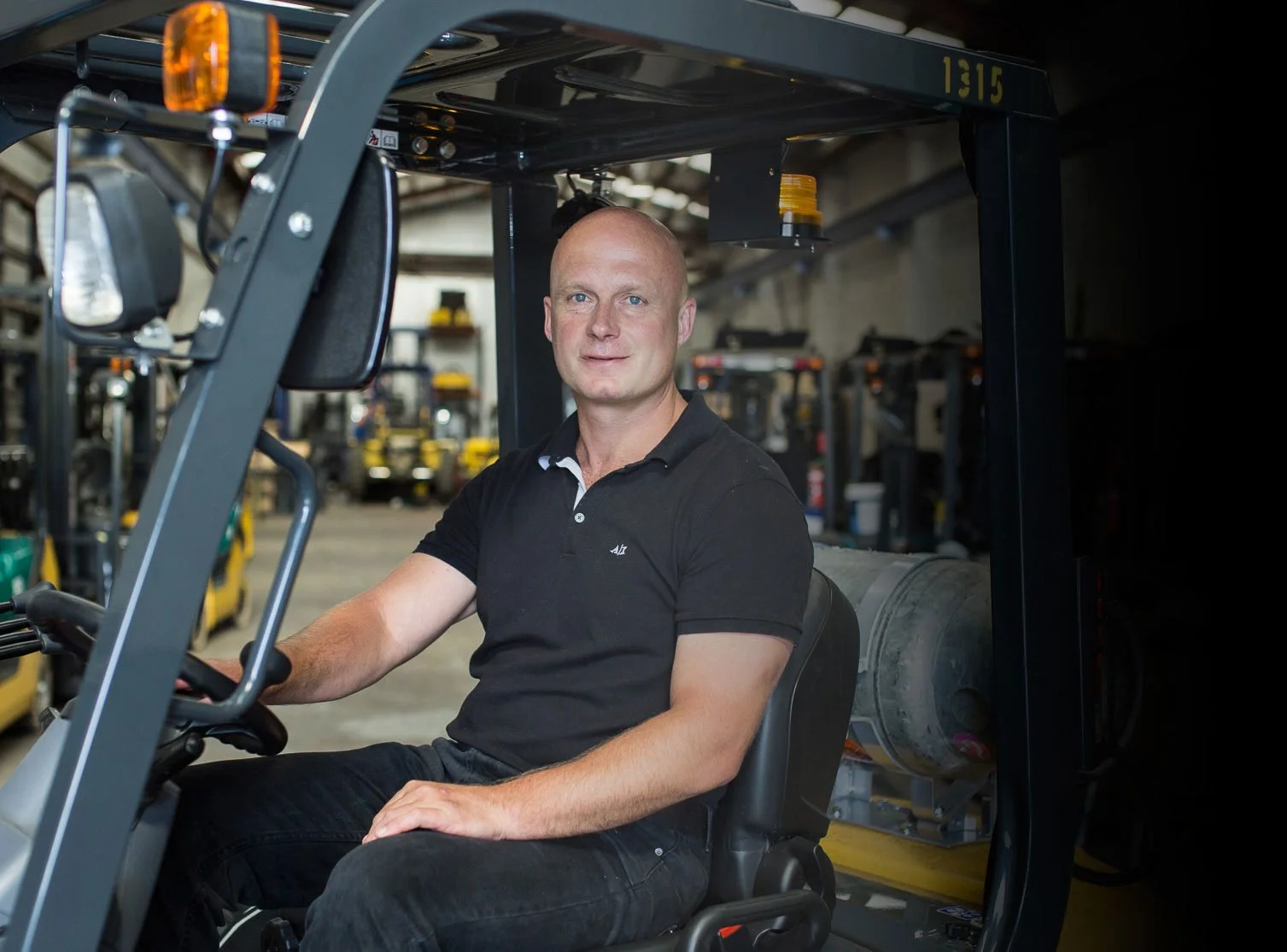 A man with a shaved head and wearing a black polo shirt sitting on a forklift inside a warehouse with shelves and multiple forklifts in the background.