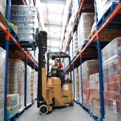 A person operating a forklift in a warehouse aisle surrounded by shelves stocked with large pallets of goods.