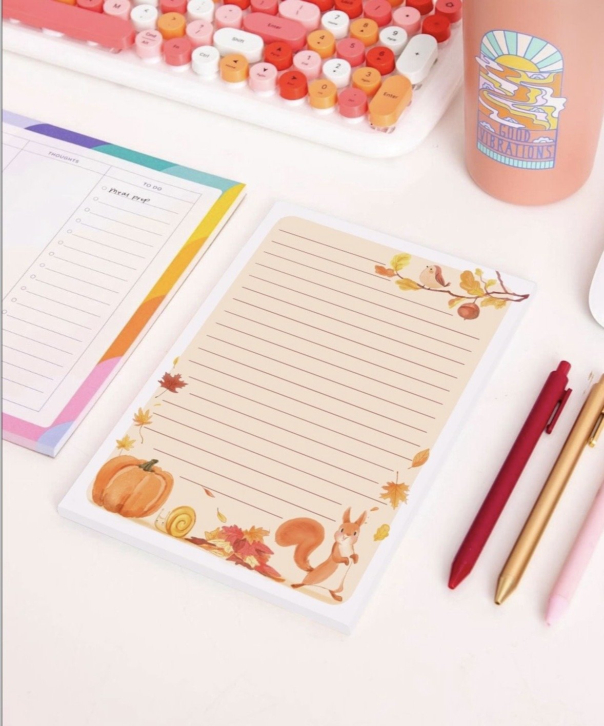 Autumn-themed stationery with lined paper decorated with a squirrel, pumpkin, leaves, and a snail. There is a notepad for a to-do list, colorful pens, a pastel-colored keyboard, a pink drink container, and a pink pen on a white desk.
