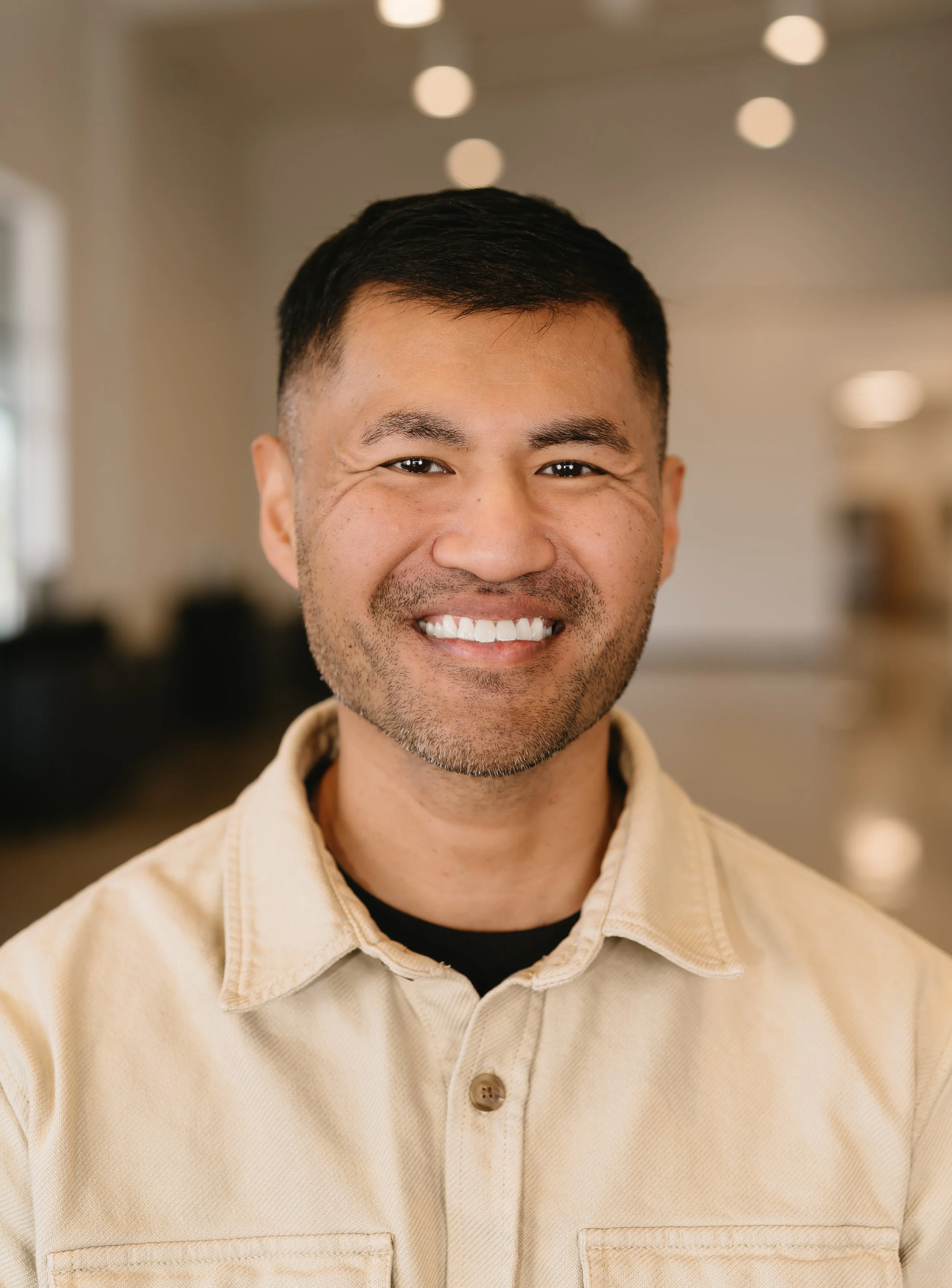 Close-up of a man with short black hair, smiling, wearing a beige jacket with a black shirt underneath, in an indoor setting with blurred lights in the background.