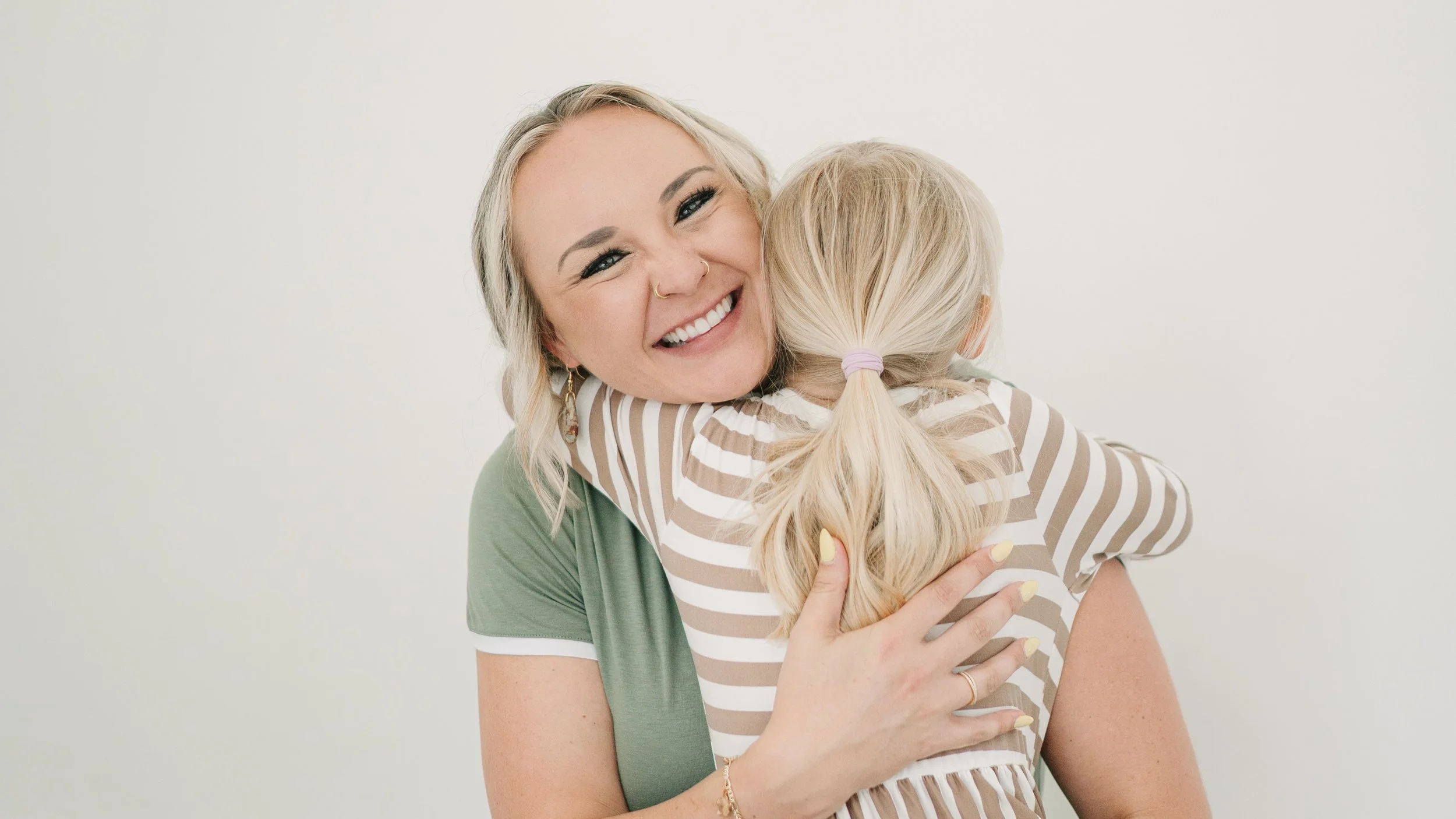 A woman with blonde hair and a nose piercing hugging a young girl with blonde hair tied in a ponytail, both smiling warmly.