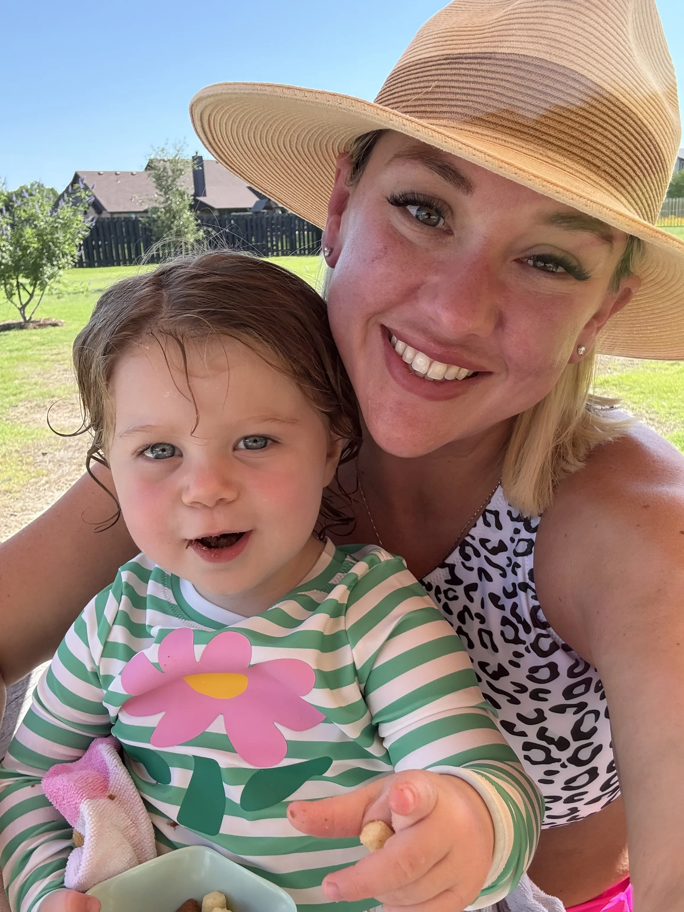 A smiling woman in a large straw hat and leopard print top, with a young girl in a striped pink and green shirt with a flower design, sitting outdoors in a backyard with trees and a house in the background. They are sharing snacks, with the woman holding a small piece of food.