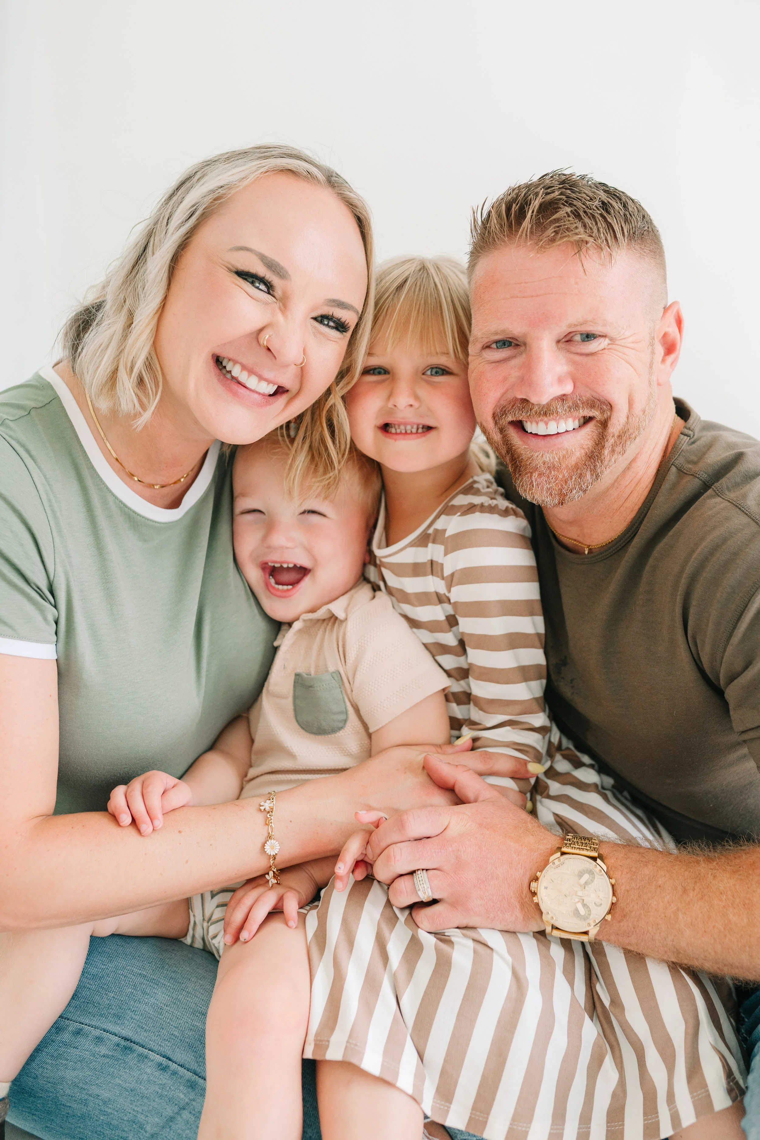 A happy family of five, including two women, two children, and a man, smiling and hugging each other in a bright room.