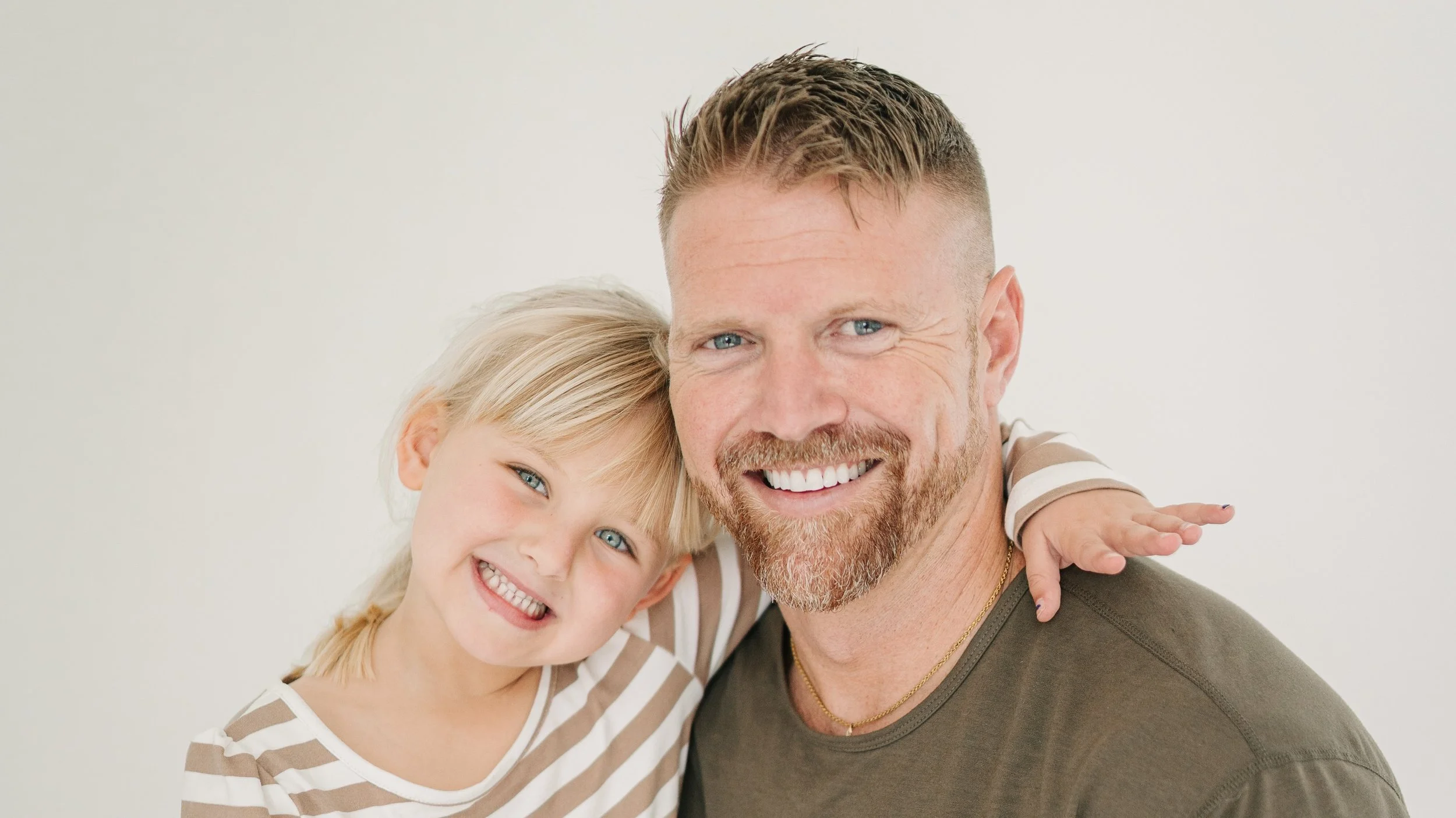A smiling man with a beard and a young girl with blonde hair and blue eyes smiling, hugging together against a plain light background.