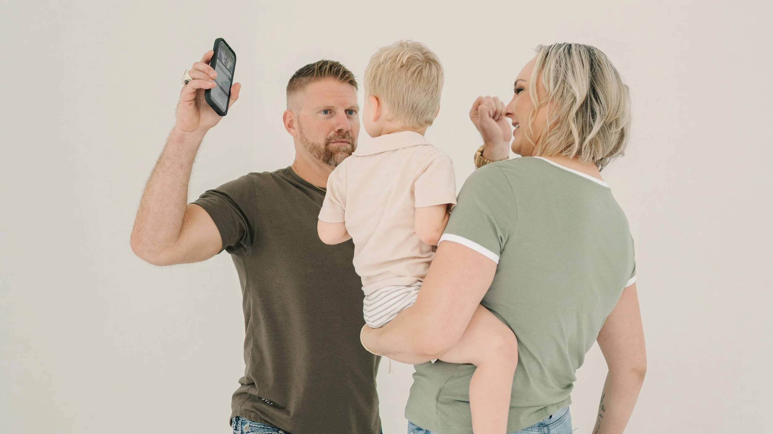 A man with a beard holding a child and a woman holding a child, with the man taking a photo of the woman and the child.
